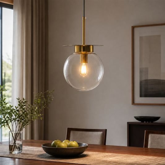 A modern pendant light with a clear glass globe shade and a brushed brass rod hanging over a wooden dining table in a tastefully decorated room.