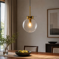 A modern pendant light with a clear glass globe shade and a brushed brass rod hanging over a wooden dining table in a tastefully decorated room.