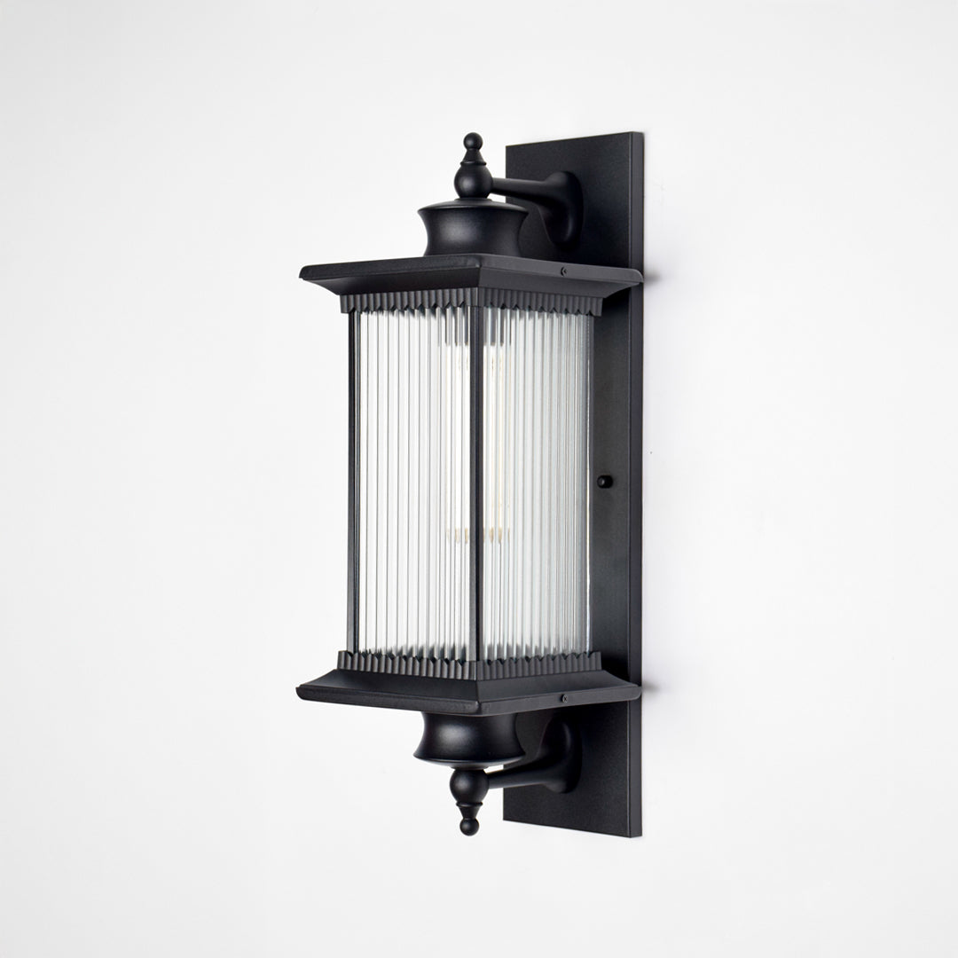 Black aluminum outdoor wall light on white background with a vertical reeded glass shade for modern patio decor.