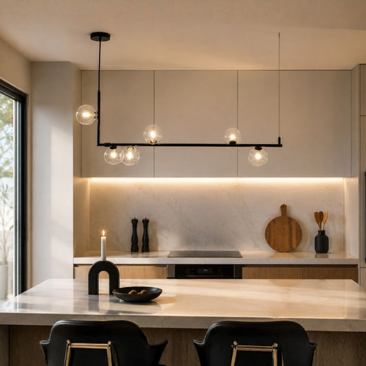 Modern linear pendant light with six clear glass globe bulbs on a black frame, hanging in a contemporary kitchen above a marble island.