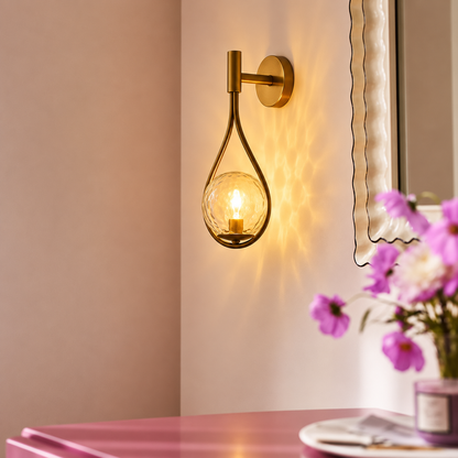 A warm brass wall sconce with a teardrop-shaped clear glass shade, mounted on a beige wall and casting a soft glow. A pink table with purple flowers is visible in the foreground.