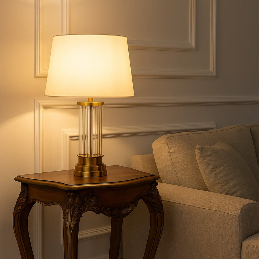 White shade gold table lamp on carved wooden side table with light on beside beige sofa for warm classic home décor.