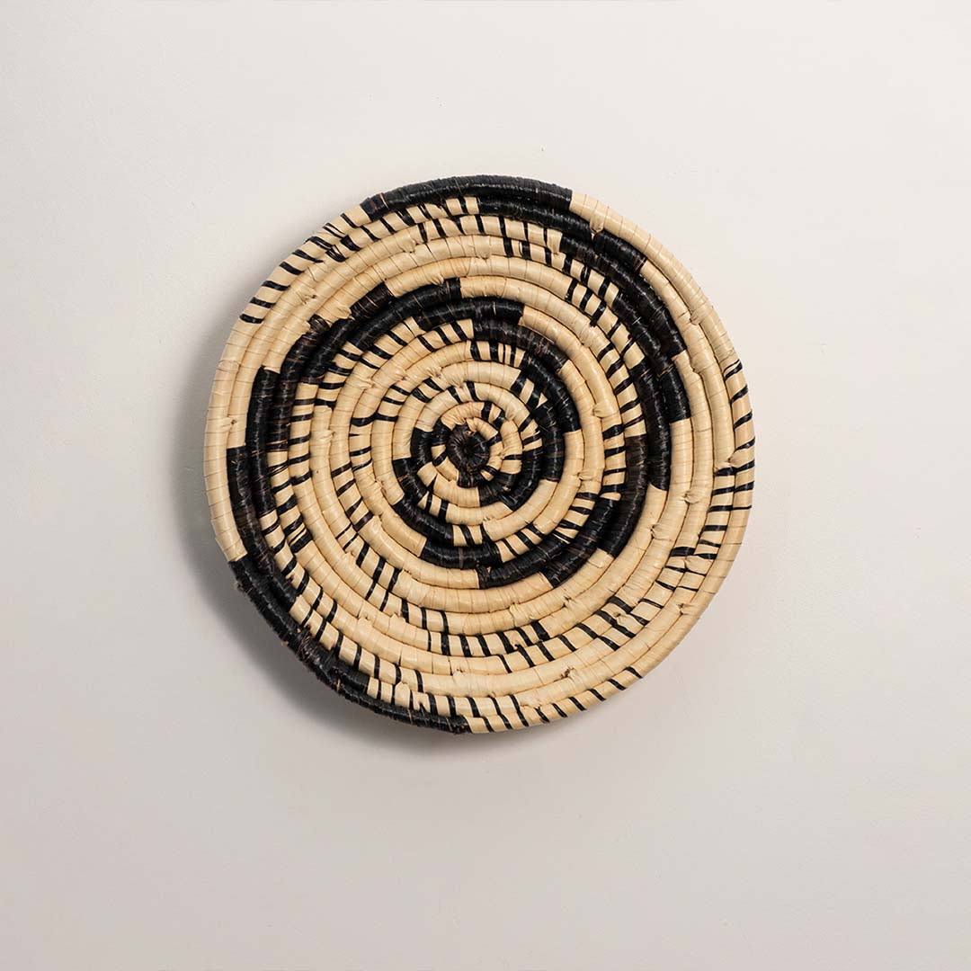 Round handcrafted black and cream woven seagrass wall basket with swirling vortex spiral pattern on white background image.