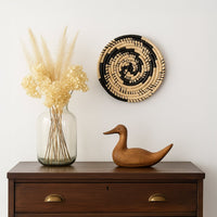 A handwoven black and natural rattan wall decor plate styled above a wooden chest with a glass vase and wooden duck creating warm modern home decor vibes.