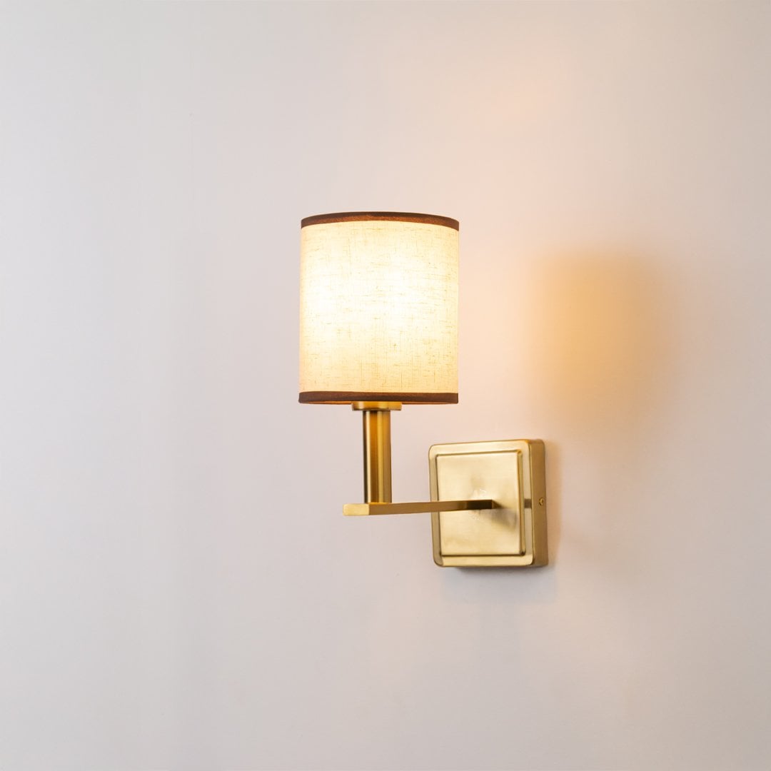 Gold wall sconce with beige fabric drum shade glowing softly on a clean white background for modern home lighting decor.