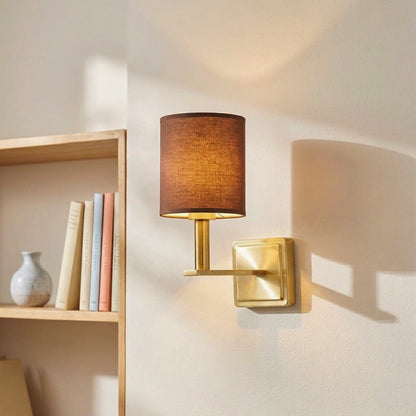 Modern gold metal wall sconce with a dark brown fabric cylinder shade casting warm light beside a wooden bookshelf.