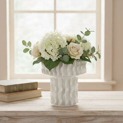 White ceramic frilled decorative vase with floral arrangement placed near window creating elegant and timeless interior decor.