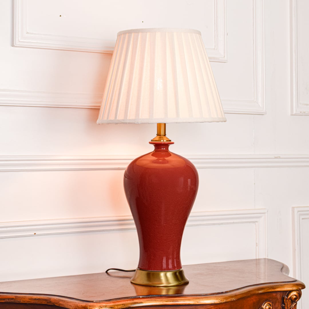 Red ceramic table lamp with white pleated shade and gold accents casting warm ambient light for elegant home decor.