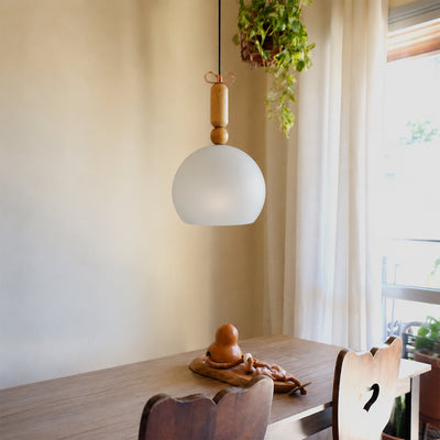 A modern pendant light with a frosted white globe shade and natural wood accents hangs over a wooden dining table in a softly lit room with a hanging plant and window curtains.