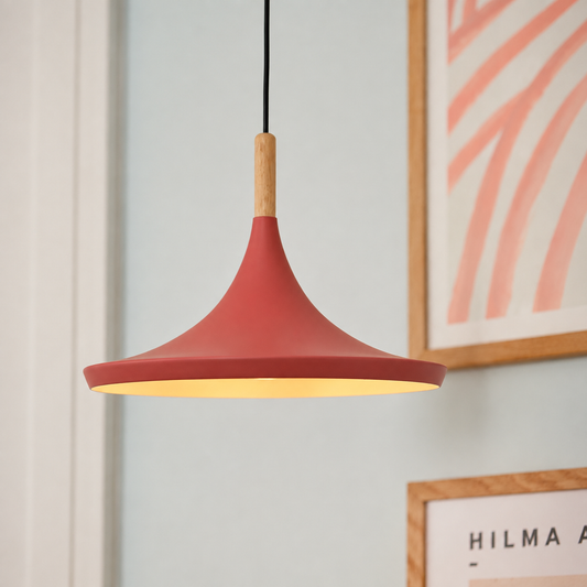 A modern pendant light with a matte terracotta red conical shade, a light natural wood neck, and a warm yellow interior glow, hanging against a light gray wall with framed art in the background.