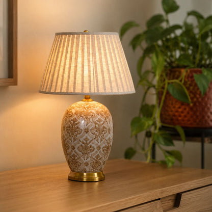 A table lamp with a patterned ceramic base and a pleated fabric shade, illuminated on a wooden console table with a blurred potted plant in the background.