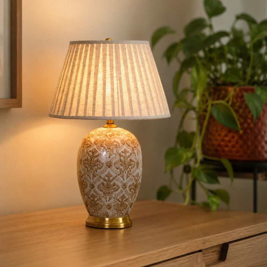 A table lamp with a patterned ceramic base and a pleated fabric shade, illuminated on a wooden console table with a blurred potted plant in the background.