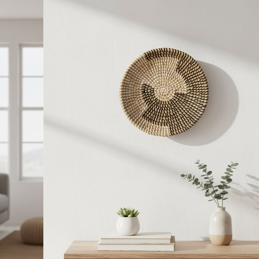 A woven wall basket hangs above a wooden shelf holding books and a vase with greenery while sunlight casts soft shadows on the white wall.