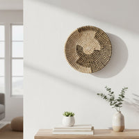 A woven wall basket hangs above a wooden shelf holding books and a vase with greenery while sunlight casts soft shadows on the white wall.