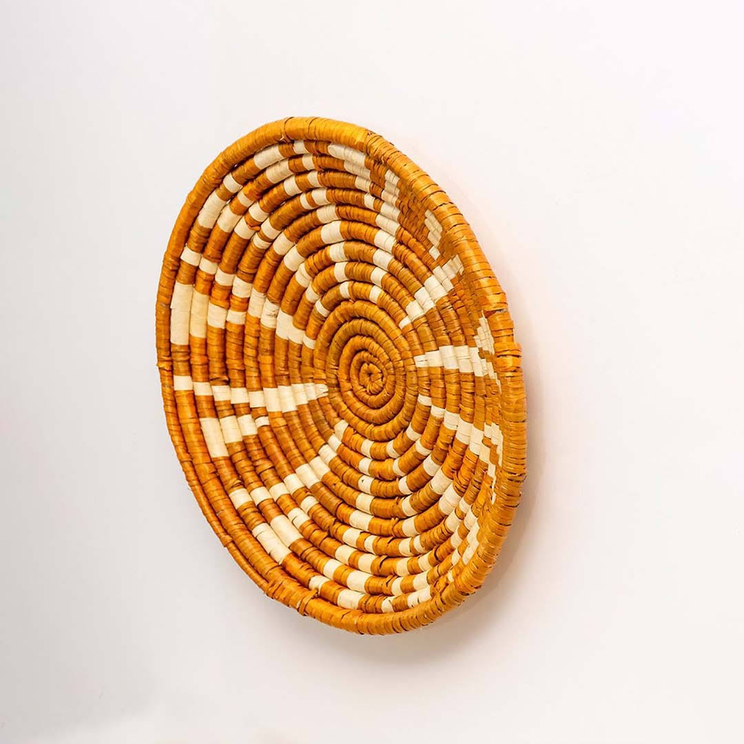 Handwoven seagrass round wall basket decor in natural beige and terracotta with raised sunburst woven profile on white.