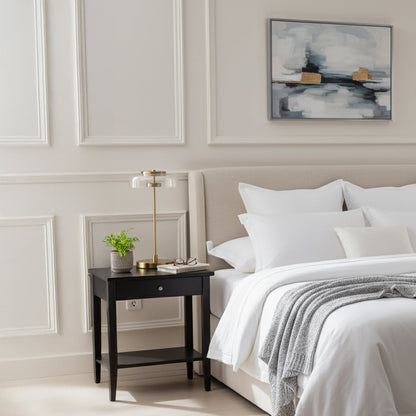 Gold and glass table lamp with a clear dome shade placed on a black bedside table creating soft warm lighting in a bright modern bedroom with white bedding.