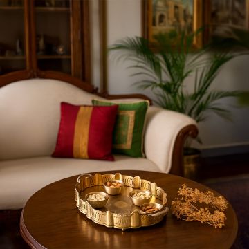 A gold decorative serving tray with matching bowls filled with assorted dry fruits displayed on a wooden table in a cozy living room with cushions and plants in the background.