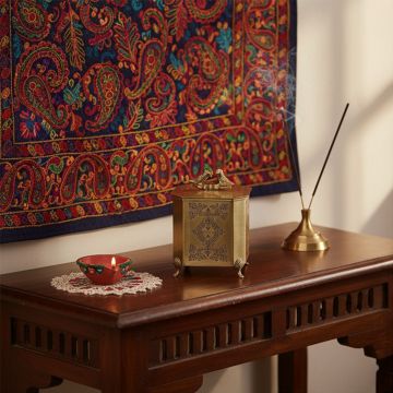 A brass decor box with bird handle placed on a wooden table beside a lit diya and incense bringing a warm traditional touch to an artistic home setting.