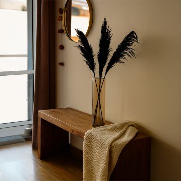 Black pampas grass styled in tall glass vase on wooden bench with cozy throw for modern home decor.