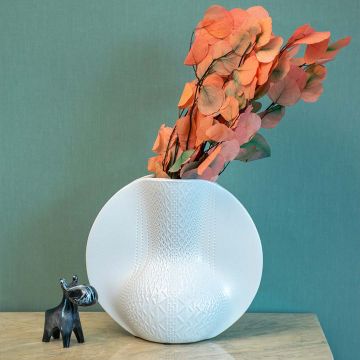Round white pottery vase with geometric pattern placed on a stone surface against a teal wall for contemporary home decor styling.