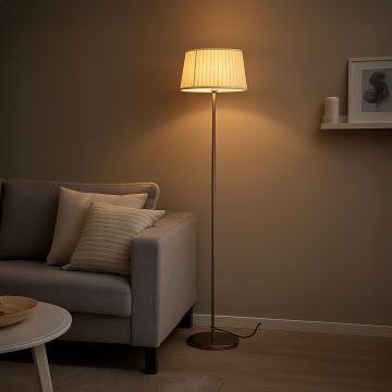 Steel floor lamp with cream pleated fabric shade beside grey sofa in modern living room warm interior lighting.