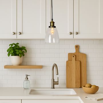 Clear glass pendant light with brass and black holder glowing warmly above modern white kitchen sink decor.