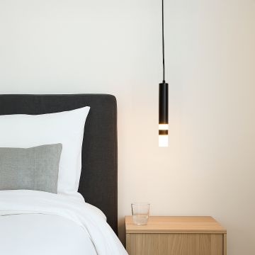 Modern black cylindrical pendant light glowing warmly beside bed with wooden side table styled in minimal bedroom interior.