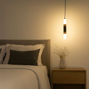 Elegant black and gold cylindrical pendant light glowing warmly beside bed with modern wooden side table and vase decor.