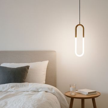 Modern gold U shaped LED pendant light glowing warmly in bedroom interior above bedside table styled with books and glass décor.