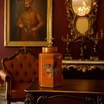 Orange ceramic decorative jar with gold horse motif and lid accent placed on wooden table in royal home interior