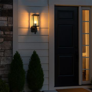 Modern outdoor wall light in black metal with glass shade glowing warmly beside front door for home entrance lighting.