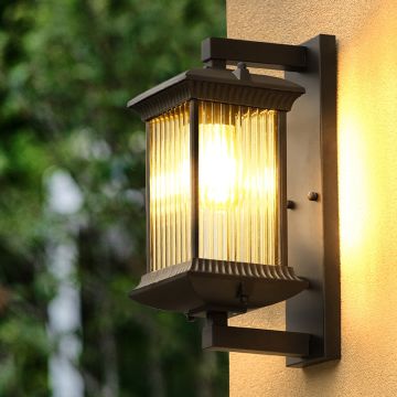 A modern outdoor wall lantern in a bronze finish mounted beside lush greenery creating a warm ambient glow that enhances the exterior space beautifully.