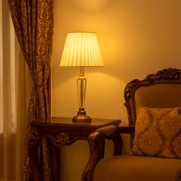 Crystal and gold table lamp with pleated shade turned on styled on wooden side table in classic bedroom decor table lamp.