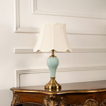 Classic table lamp with beige fabric shade and light blue ceramic base with gold finish placed on a carved wooden console against white panel wall.