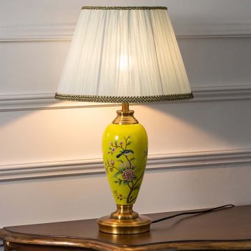 Yellow ceramic table lamp with hand-painted floral design and pleated white shade turned on against white background radiating soft warm light.