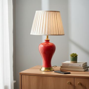 Red ceramic table lamp with glossy curved body gold metal base and pleated fabric shade styled on bedside table.