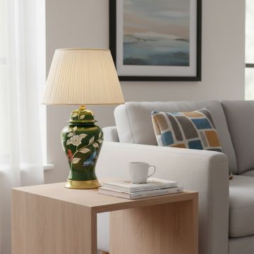 Green ceramic table lamp with hand painted floral butterfly artwork gold metal base and pleated fabric shade styled on side table.