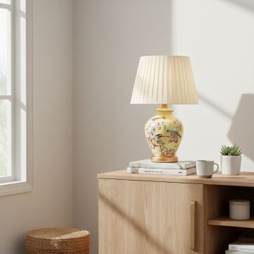 Yellow ceramic table lamp with hand painted floral bird artwork gold metal base and pleated fabric shade styled on side table.