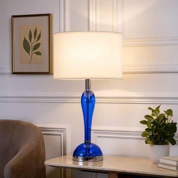 Blue glass lamp with white fabric shade glowing warmly placed on a side table with wall art plant and sofa in living room.