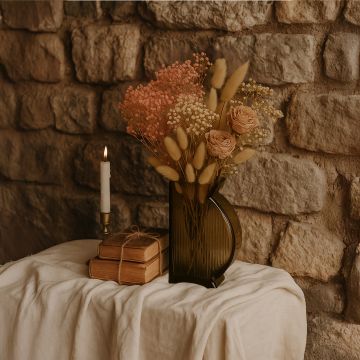 Smoky glass vase styled with roses and dried florals on rustic table against stone wall creating warm vintage decor.