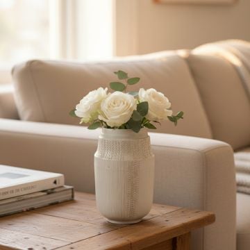 White ceramic small vase with white roses placed beside sofa creating warm and elegant living space decor.