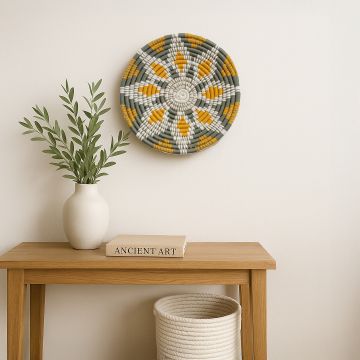 Handcrafted woven wall plate with green mustard and cream patterns displayed on a white background above a wooden console styled with books a ceramic vase and minimal decor for a calm lifestyle scene.