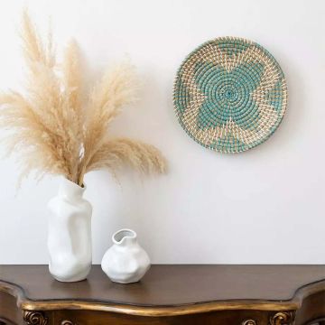 Handwoven jute wall basket decor in beige with turquoise spiral pattern styled on wooden console for rustic boho living room.