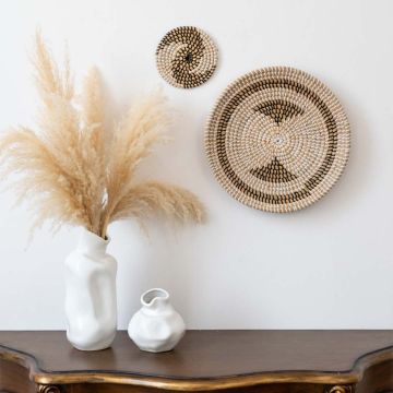 Handwoven round wall plate in natural beige and black pattern styled with white vases for home decor.