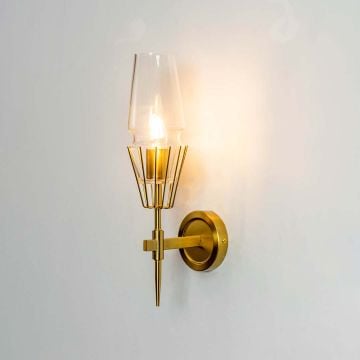 Illuminated gold brass wall lamp with clear tapered glass shade isolated on white background highlighting warm bulb glow for interior lighting.