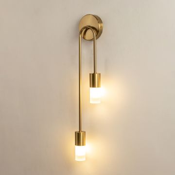 A gold metal dual wall sconce with frosted glass cylinder shades shown with lights on against a white background highlighting its warm soft illumination.