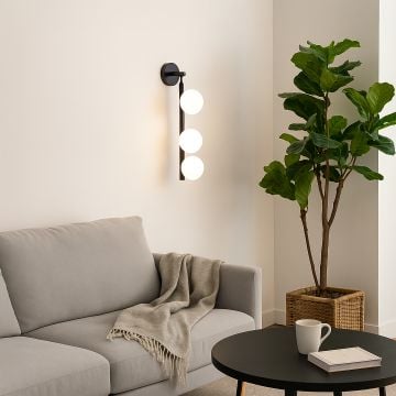 Modern black wall sconce with three frosted glass globes glowing warmly in living room beside sofa and indoor plant for cozy décor.