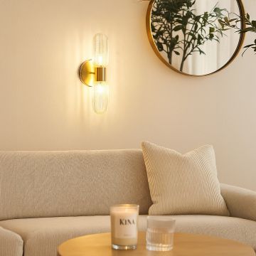 Elegant gold wall sconce with dual textured glass tube shades glowing warmly in living room interior beside sofa and mirror décor.