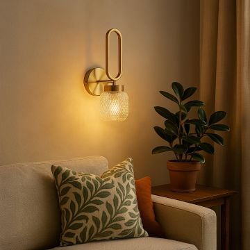 Elegant gold wall sconce with loop design and textured glass shade glowing warmly in living room beside sofa and indoor plant.