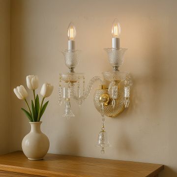 Double arm crystal wall light with gold base glowing warmly beside vase with white tulips in modern decor.